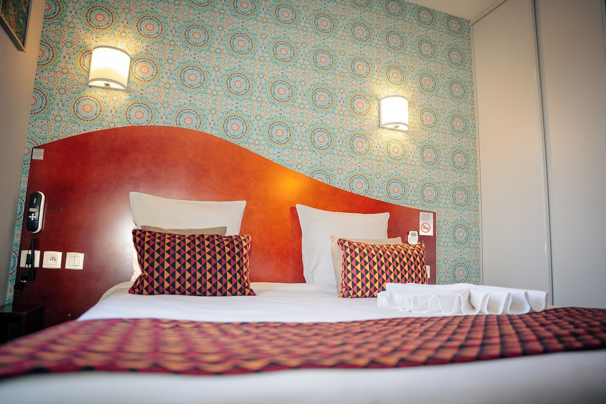 Habitación de hotel con papel tapiz estampado, cabecera roja y dos lámparas fijadas a la pared.
