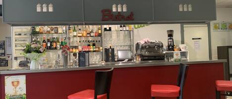 Un espace bar avec un comptoir rouge, deux tabourets de bar rouges, une machine à café et des étagères remplies de diverses bouteilles et de verres.