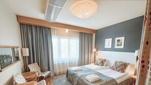 A hotel room with two beds, a desk, chairs, a large window with curtains, a ceiling fan, and wall-mounted lamps.