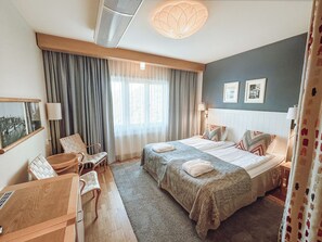 A hotel room with two beds, a desk, chairs, a large window with curtains, a ceiling fan, and wall-mounted lamps.