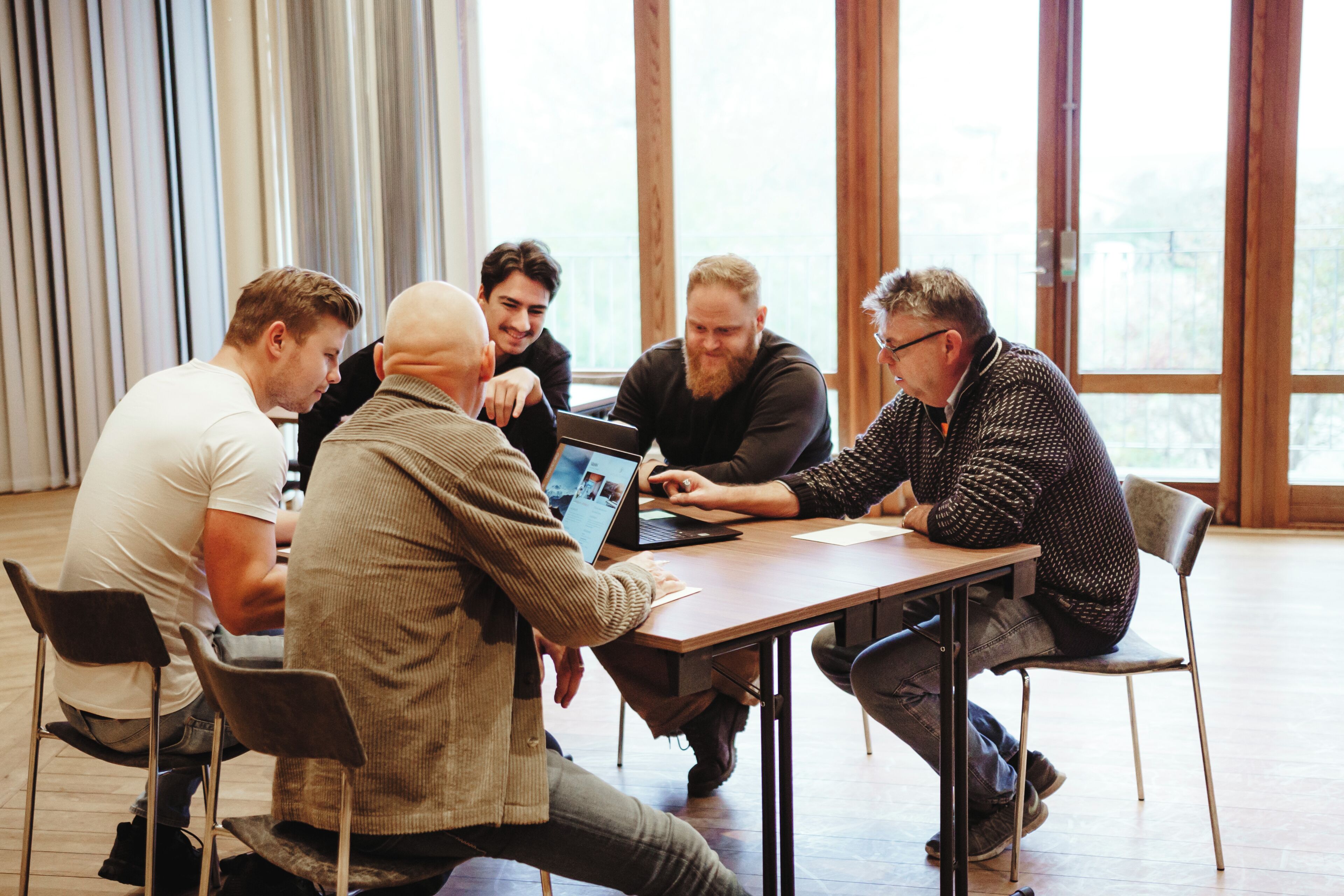 Un grupo de personas sentadas alrededor de una mesa en una habitación con amplias ventanas y piso de madera.