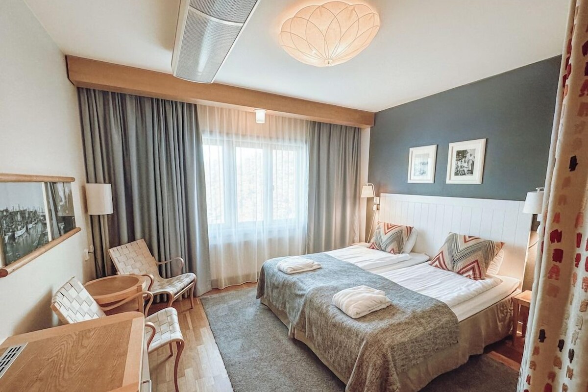 Quarto de hotel com cama, duas cadeiras, escrivaninha, janela grande com cortinas e luminária no teto.
