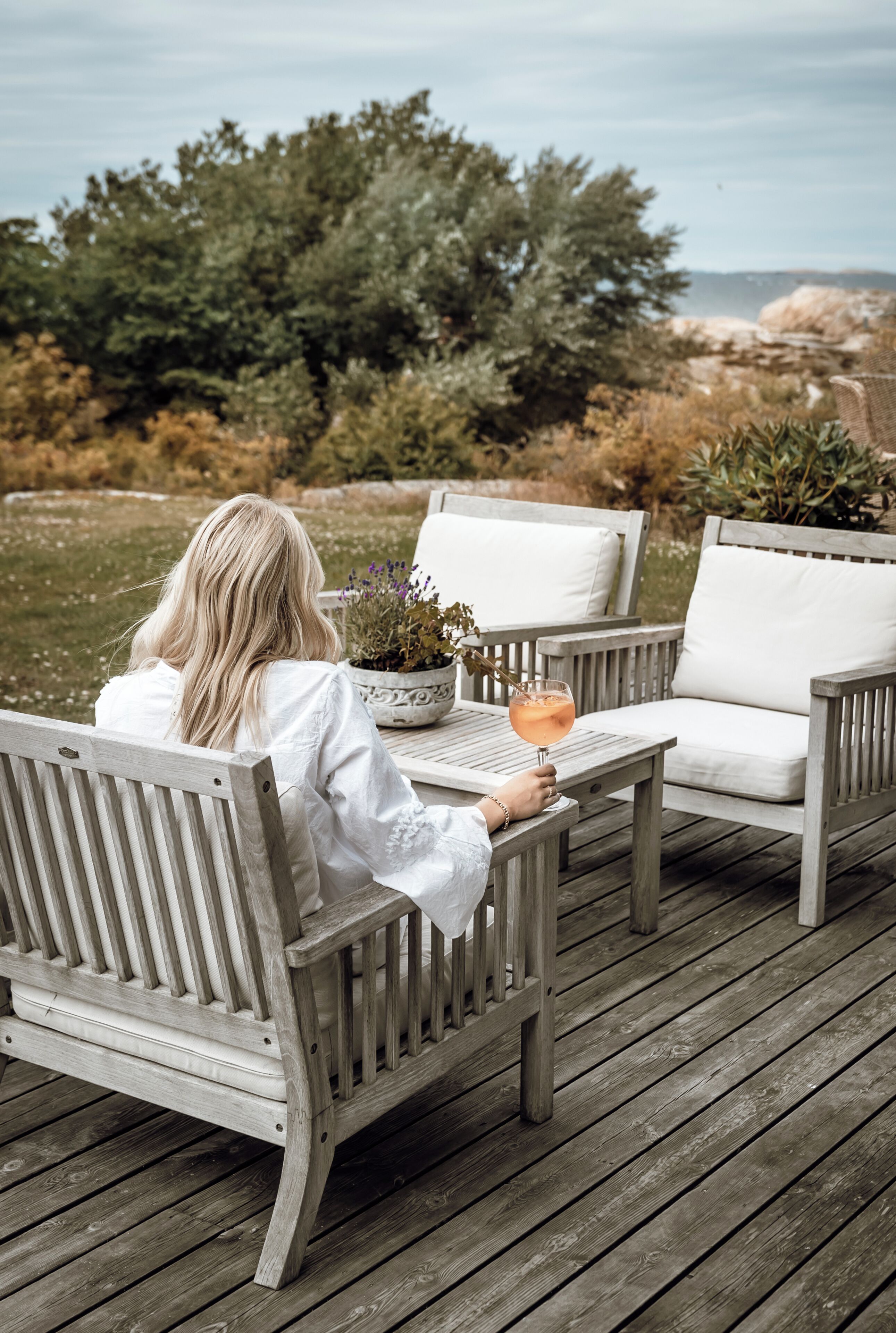 A person seated on a wooden deck chair with a white cushion, holding a drink, with a potted plant on the table.
