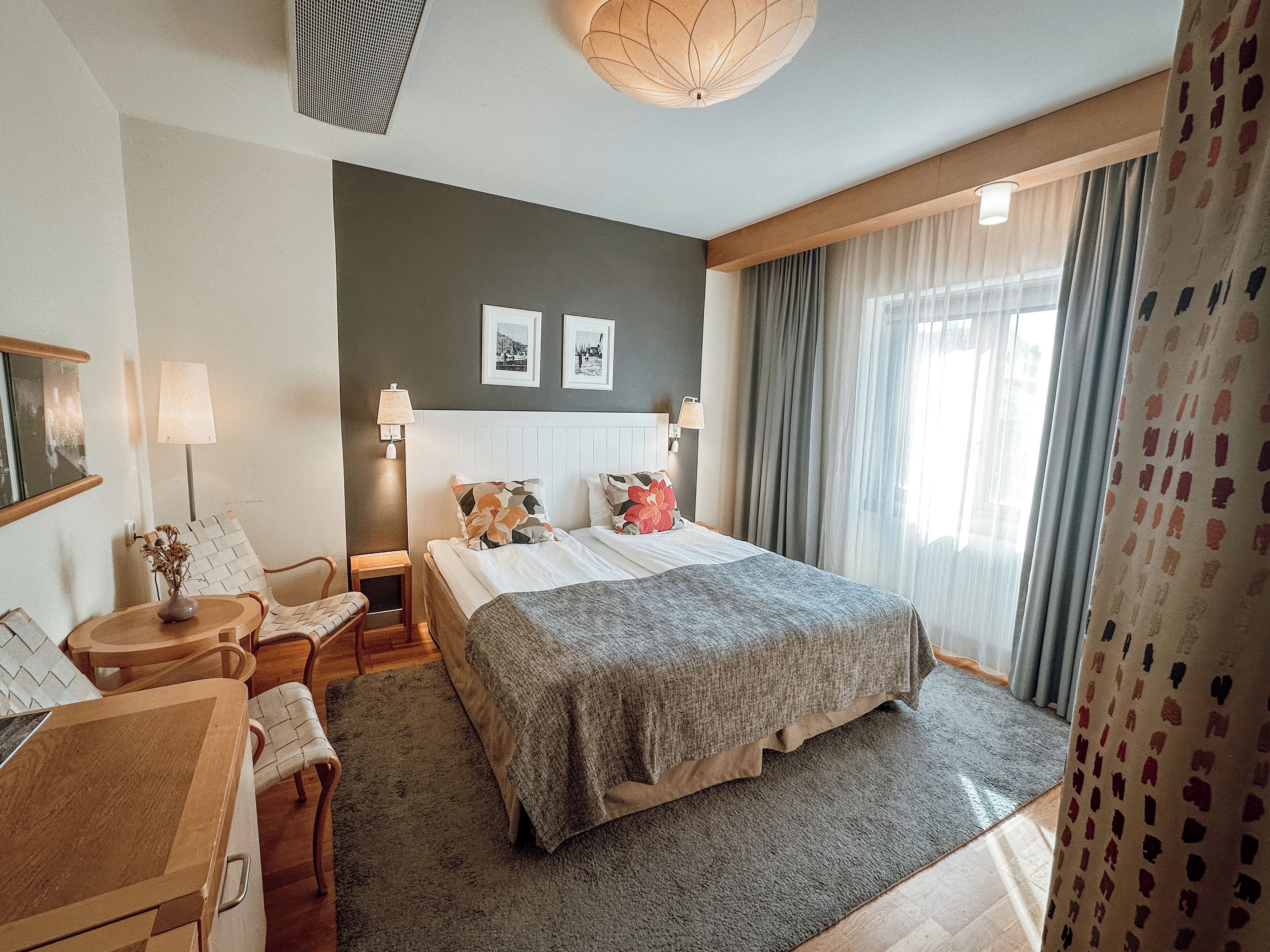 Una habitación de hotel con una cama, dos mesitas de noche, un escritorio, una silla, una lámpara, un televisor montado en la pared y una ventana con cortinas.