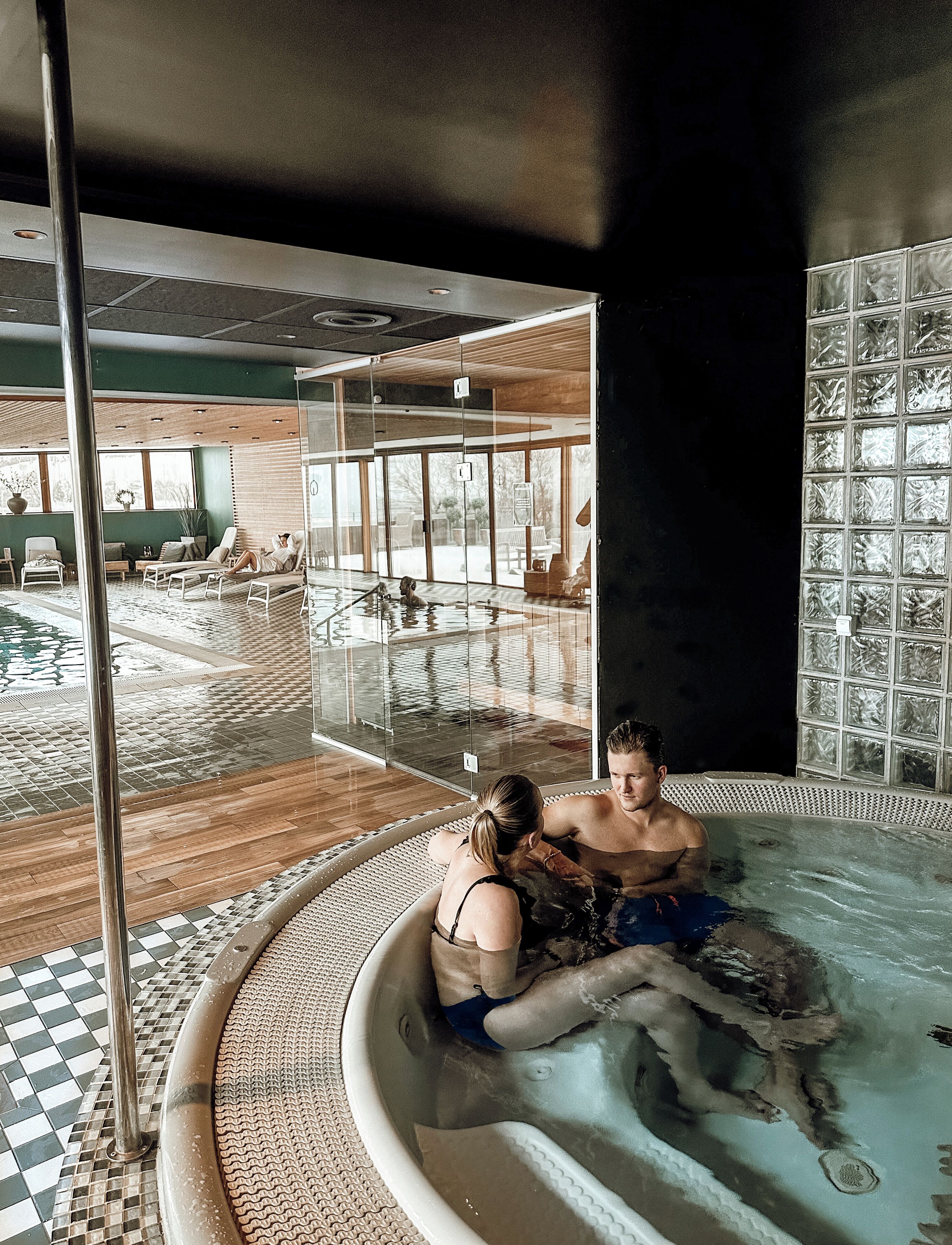 Una pareja en un jacuzzi con vistas a una piscina y un muro de cristal.