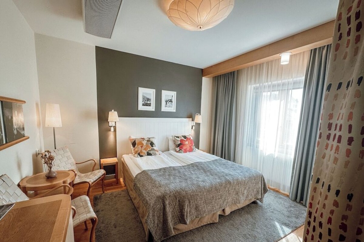 A hotel room with a bed, bedside tables, a desk, a chair, a lamp, and a large window with curtains.