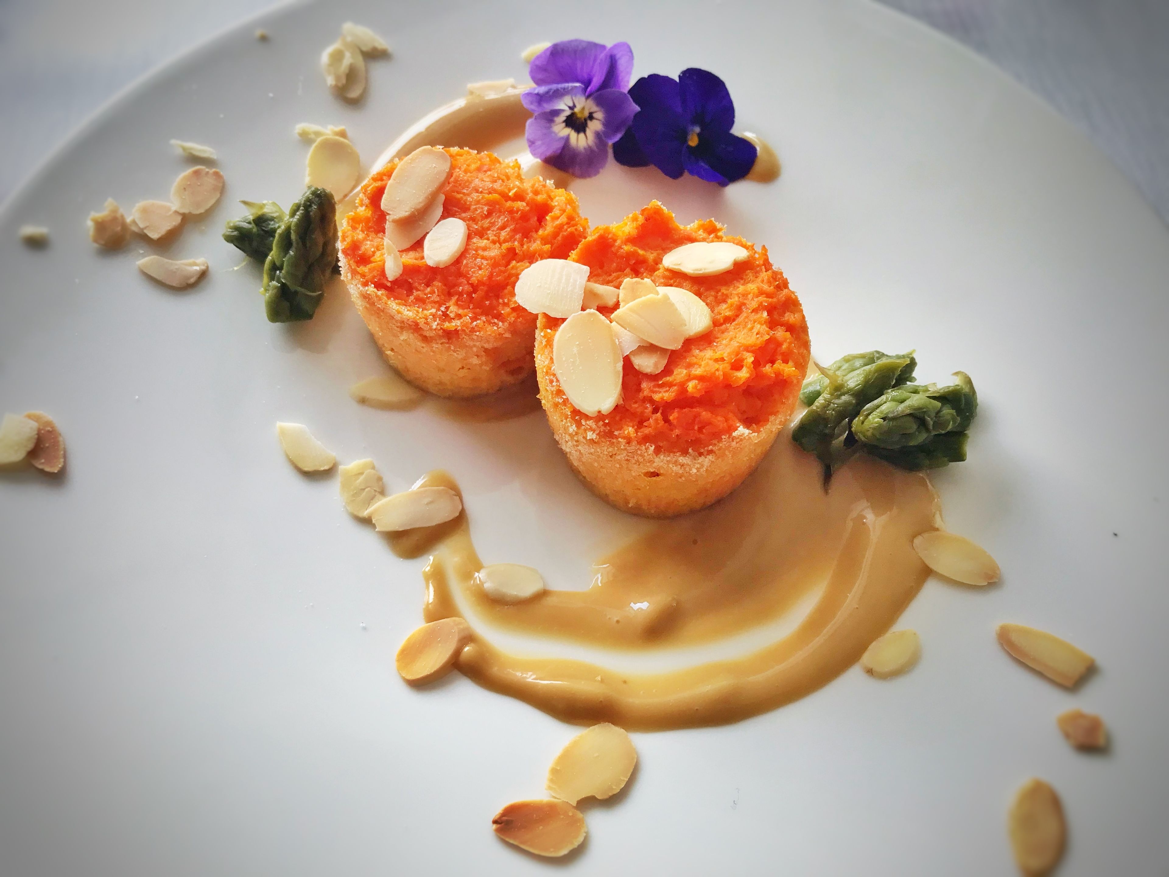 Um prato de sobremesa com dois itens arredondados de cor laranja, cobertos com amêndoas fatiadas, acompanhados de um fio de molho marrom claro e decorados com uma pequena flor roxa e folhas verdes.