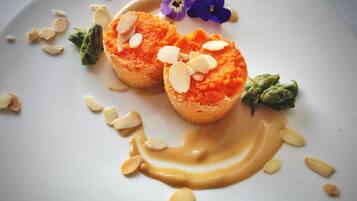 Um prato de sobremesa com dois itens arredondados de cor laranja, cobertos com amêndoas fatiadas, acompanhados de um fio de molho marrom claro e decorados com uma pequena flor roxa e folhas verdes.