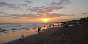 A beach with people walking and sitting, a sunset in the background, and a calm sea. - Aldeoz Grand Kancana Villa Bali (Keroboken Kelod)