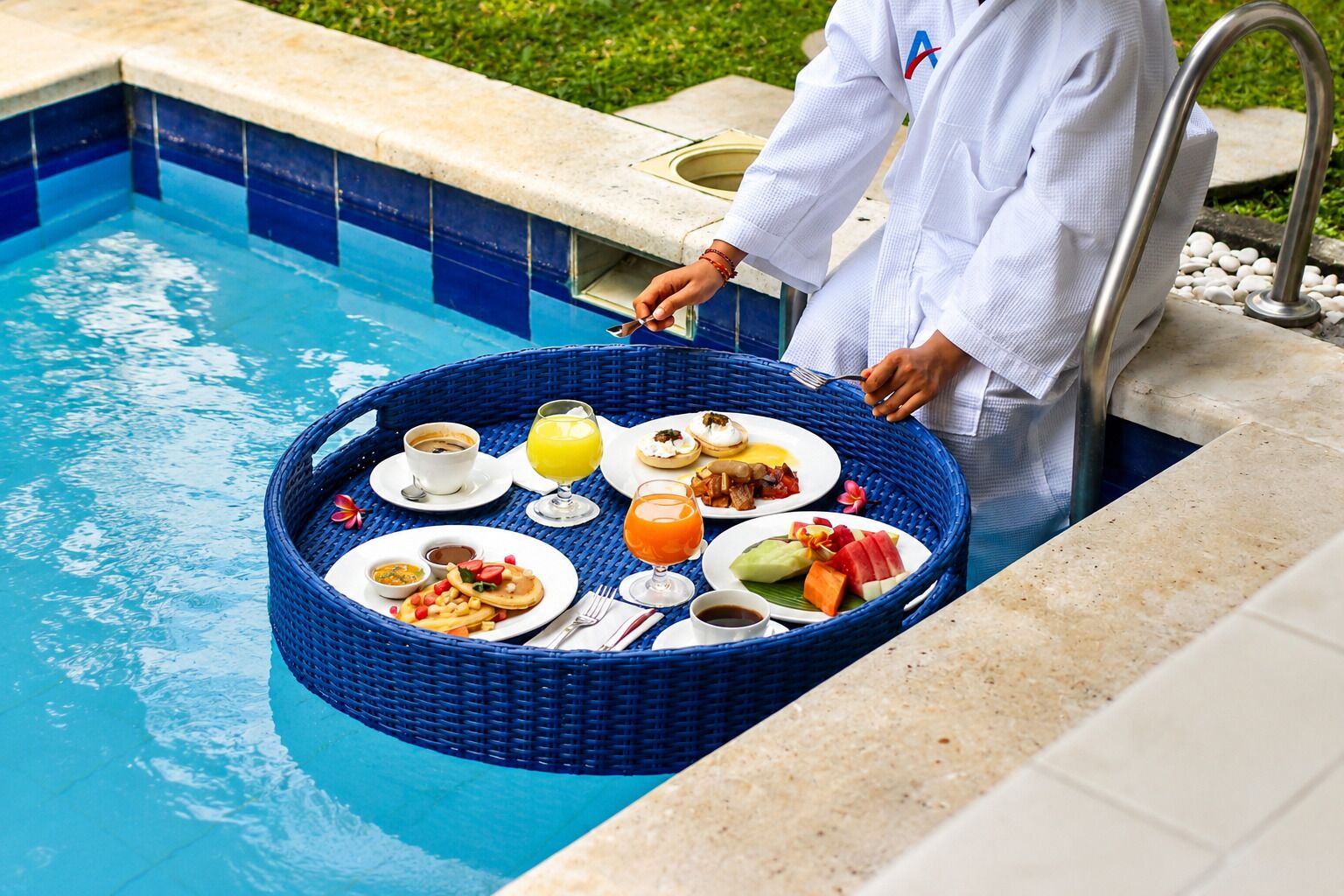 A poolside breakfast setup with a tray of food and drinks, a person in a white robe, and a pool with blue tiles.