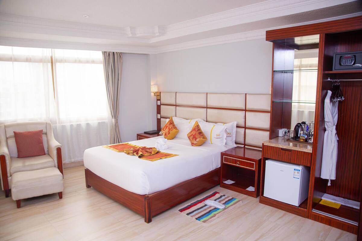 A hotel room with a large bed, a chair, a small refrigerator, a vanity with a mirror, and a window with curtains.