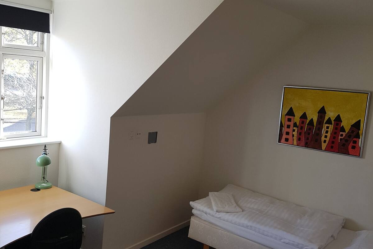 A small, well-lit room with a desk, chair, and a bed. There is a window with blinds, a ceiling light, and a wall-mounted picture.