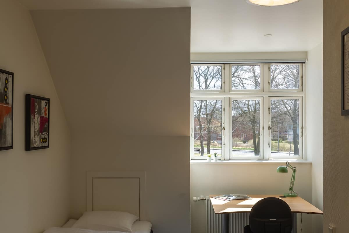 A bedroom with a bed, a desk, and a chair. There is a window with a view of trees and a building.