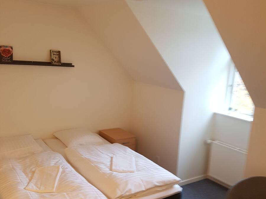 Une petite pièce bien éclairée, avec un lit, une table de chevet et une fenêtre.