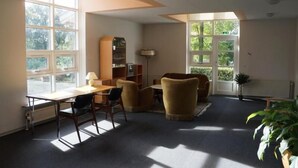 A room with a desk, two chairs, a bookshelf, and a lamp. There are large windows with a view of greenery outside. - Danhostel Brande (Brande)