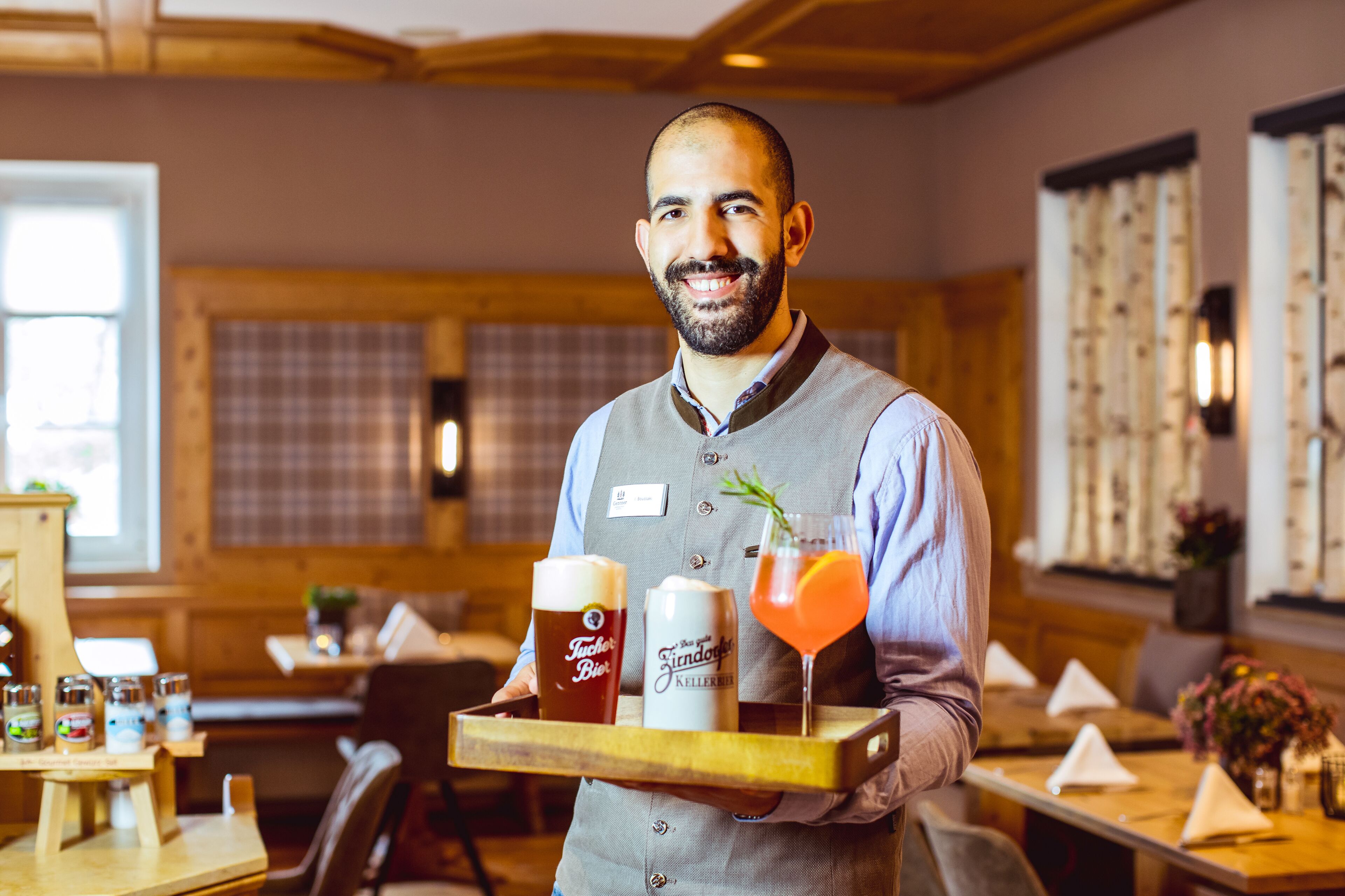 A person holding a tray with a beer mug, a can, and a cocktail glass in a restaurant setting.