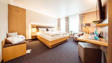 A hotel room with a large bed, a desk, a chair, a small table, a TV, and a plant.
