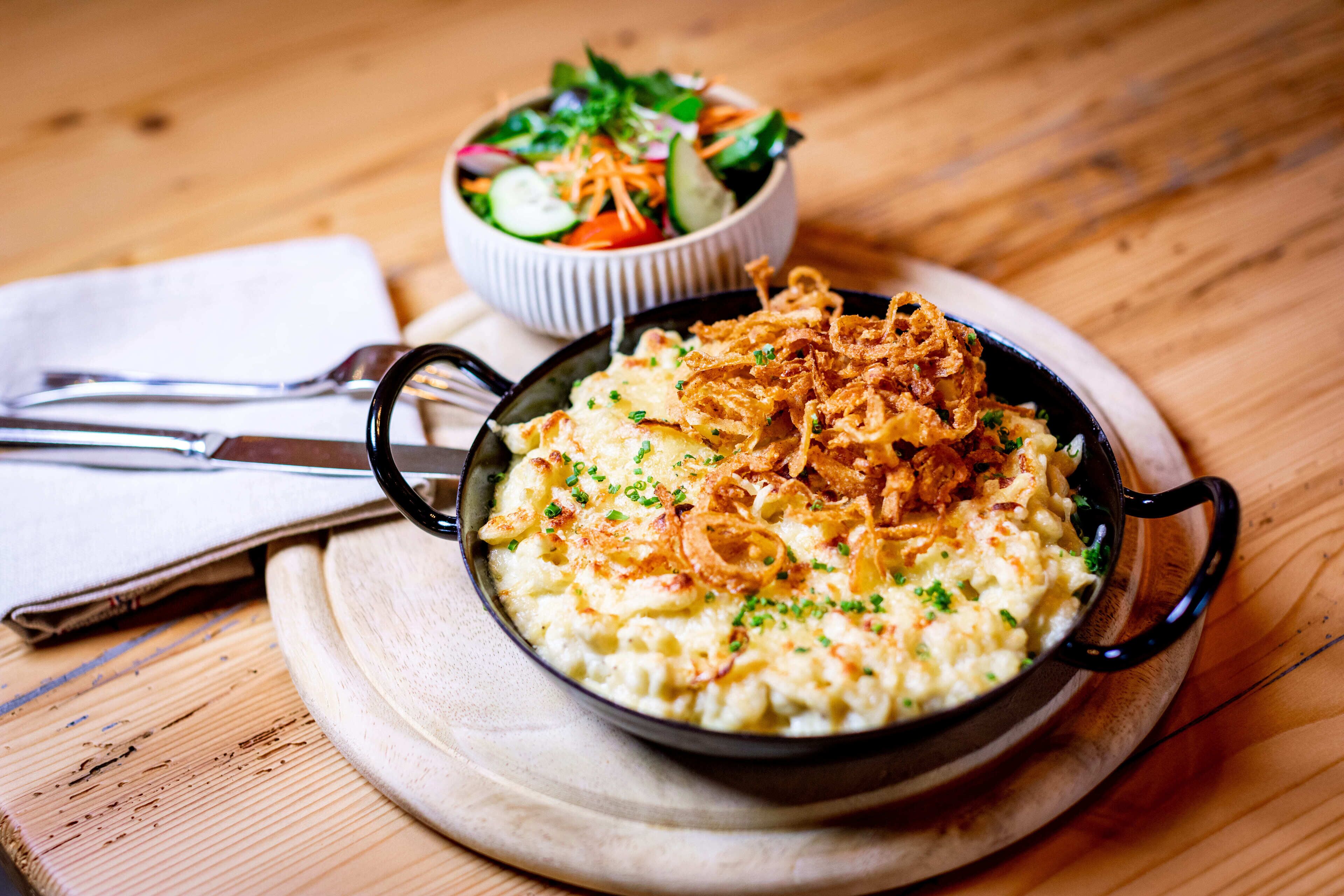 A bowl of creamy food topped with fried onions, a side salad, and a napkin with utensils on a wooden table.
