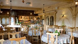 A rustic dining area with wooden tables and chairs, a bar in the background, and a stone wall with arched windows.