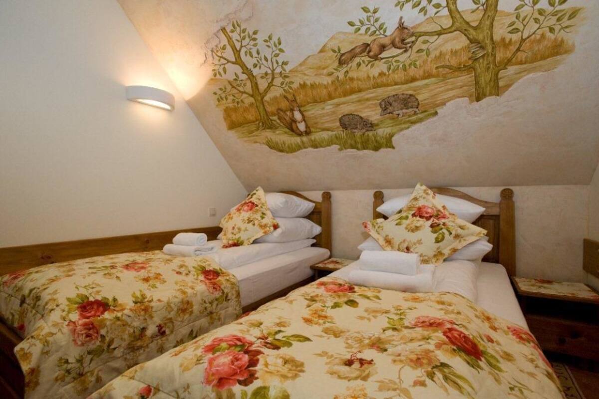Une chambre avec deux lits, chacun agrémenté de draps à motifs floraux et d’oreillers assortis. On y trouve également un luminaire fixé au mur et une fresque représentant un paysage naturel avec des animaux.