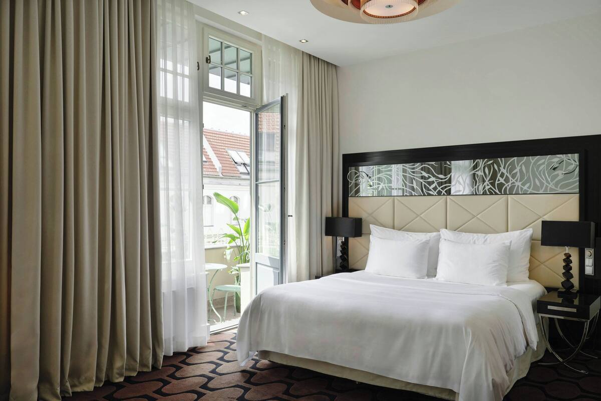 Habitación de hotel con cama, mesita de noche, lámpara y vista al exterior a través de un amplio ventanal.