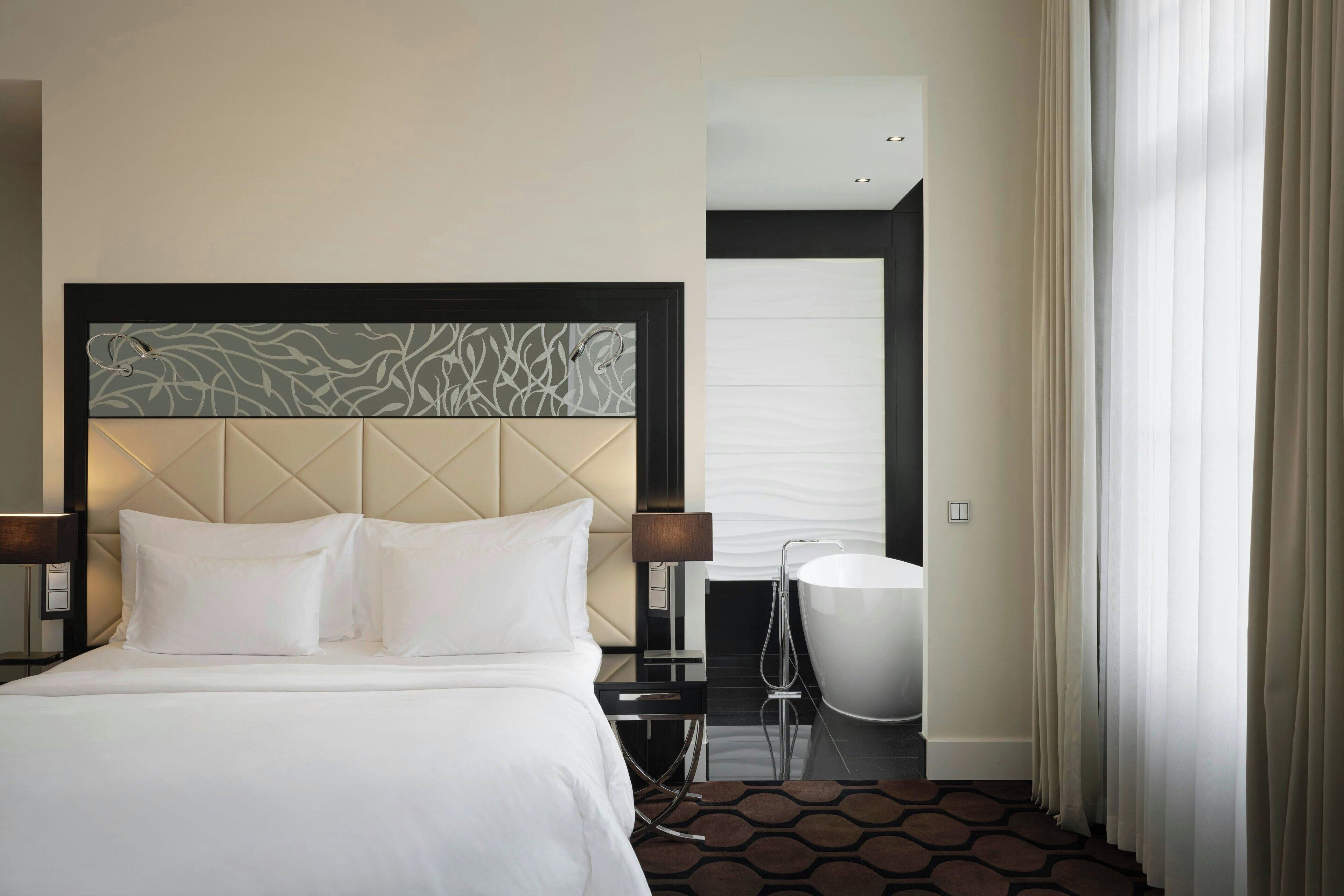 Quarto de hotel com uma cama grande, cabeceira moderna, mesa de cabeceira com abajur, uma cadeira e banheiro com banheira.