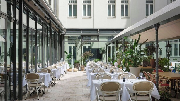 Ein großzügiger Außenbereich zum Essen mit Tischen und Stühlen, die entlang einer Glaswand angeordnet sind und einen Garten überblicken.