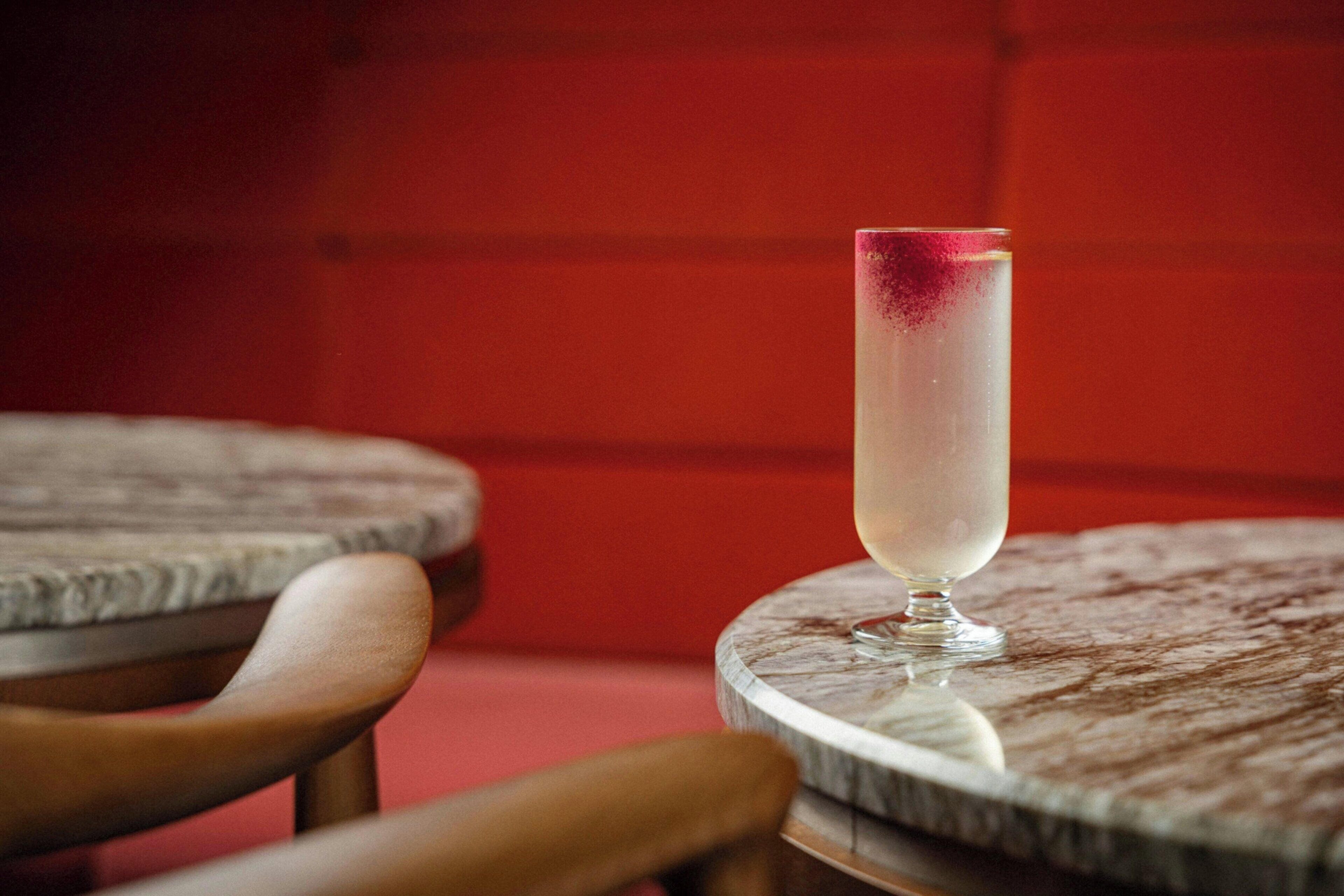 Una mesa con un vaso de una bebida transparente, posiblemente un cóctel, y una guarnición roja en la parte superior. La mesa tiene una superficie similar al mármol y está rodeada de sillas de madera.