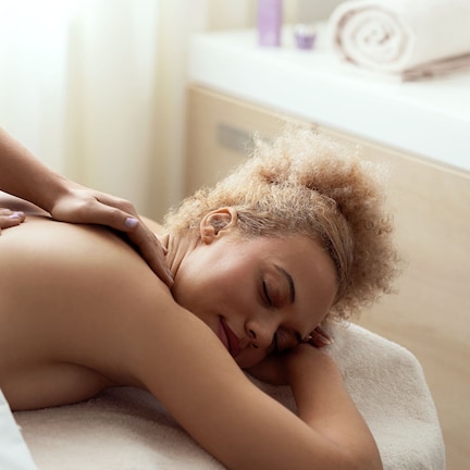 A person receiving a back massage in a room with a white towel, a wooden cabinet, and a rolled-up towel on a counter.