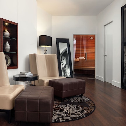 A modern living room with a sofa, ottoman, and a round rug. There is a side table with a lamp, a mirror, and a shelf with decorative items. The room has wooden flooring and a door leading to another area.
