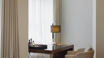 A hotel room with a curved desk, a chair, a lamp, and a large window with sheer curtains.