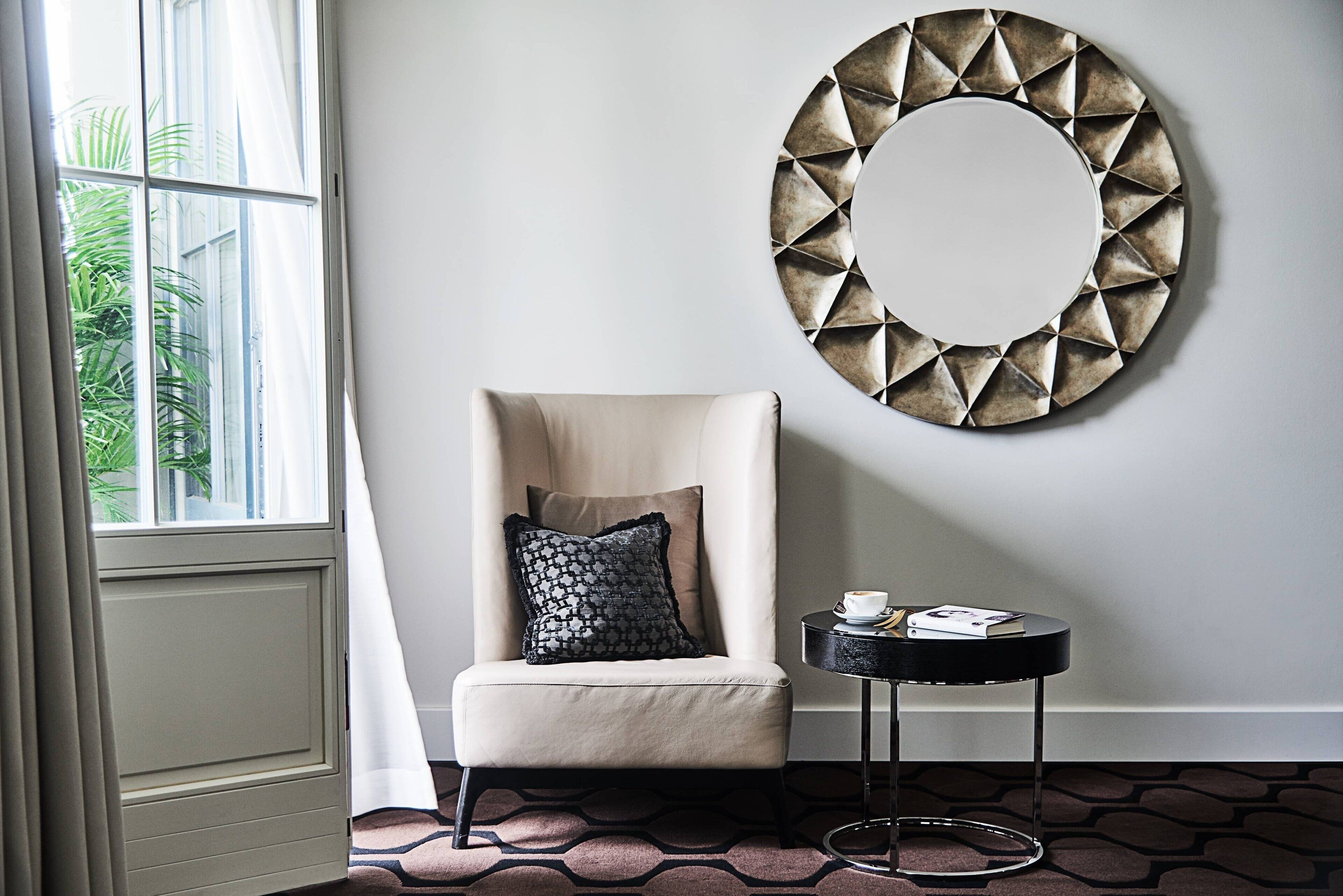 A room with a chair, a round wall decoration, a small table with a book and a cup, and a patterned carpet.