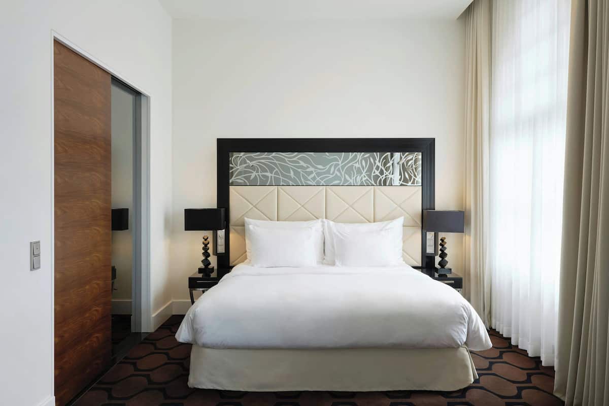 Una cama bien hecha con cabecera, dos mesitas de noche con lámparas, y vista a una ventana con cortinas.