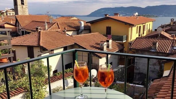 A balcony with two glasses of red drink and a bowl of snacks, overlooking a town with terracotta roofs and a tower. - Affittacamere Piazza (Verbania)