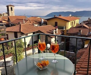 A balcony with two glasses of red drink and a bowl of snacks, overlooking a town with terracotta roofs and a tower. - Affittacamere Piazza (Verbania)