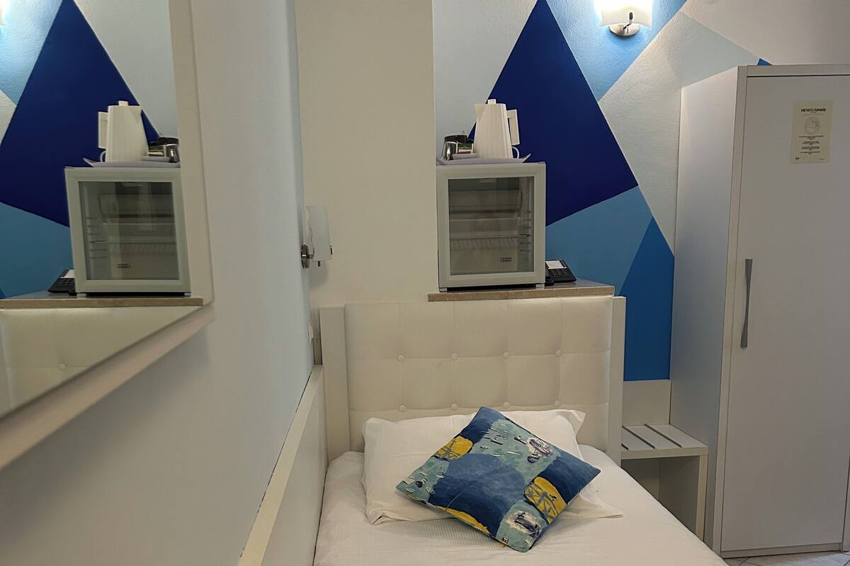 Una habitación pequeña y ordenada con cama, escritorio, silla, refrigerador y estante con un ordenador.