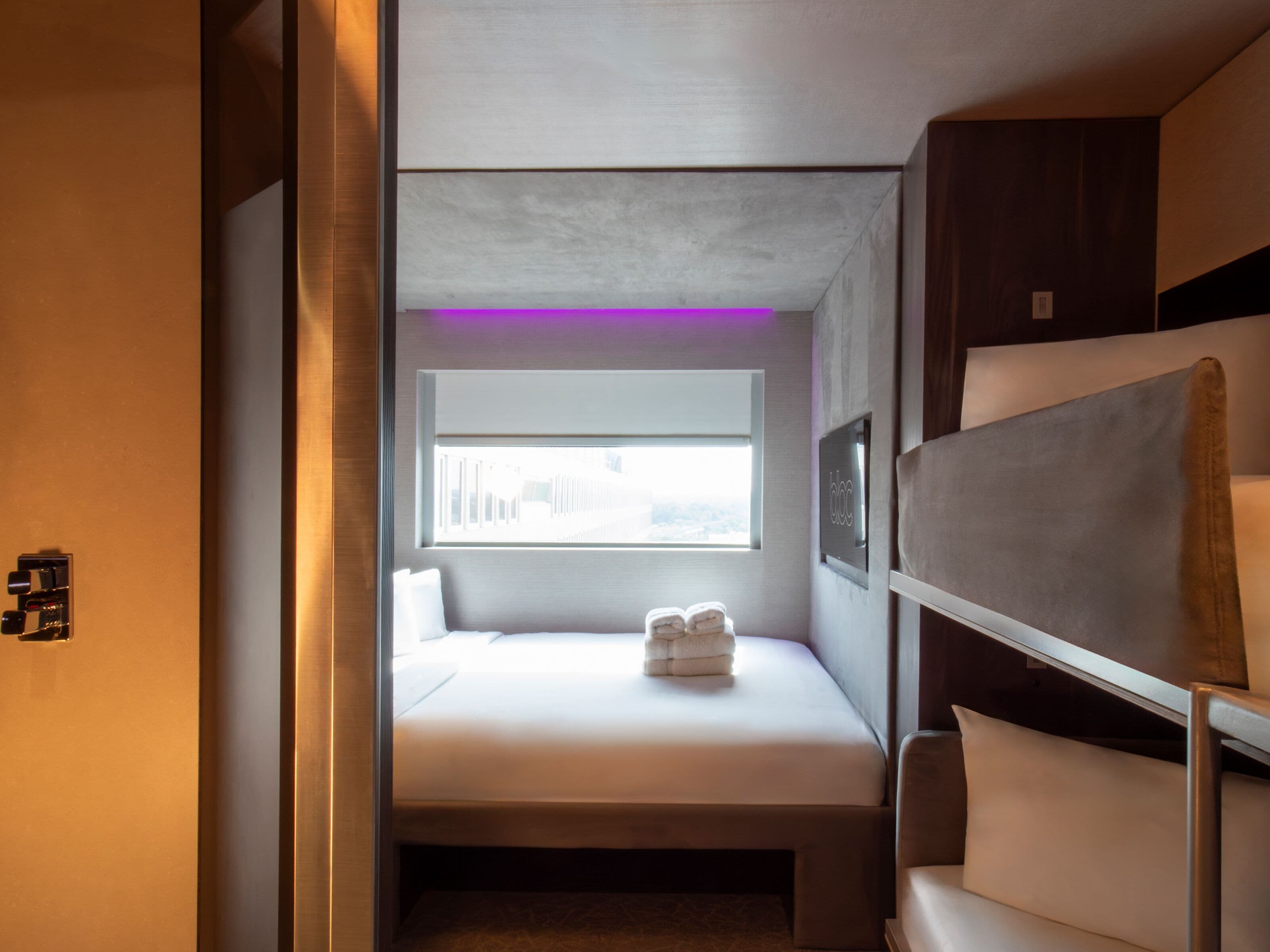 A compact, modern hotel room with a bed, a small window, and a seating area with a sofa and a chair.