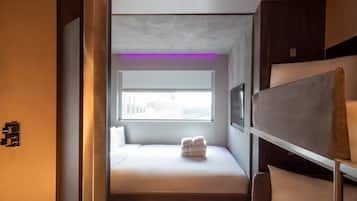 A compact, modern hotel room with a bed, a small window, and a seating area with a sofa and a chair.