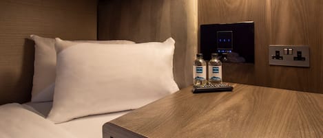 A hotel room with a wooden headboard, a bedside table with two bottles of water, a remote control, and a wall-mounted power outlet.