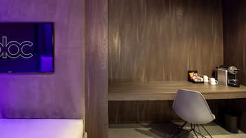 A modern hotel room with a wooden desk, a white chair, a television on the wall, and a small table with a coffee maker and a trash bin.
