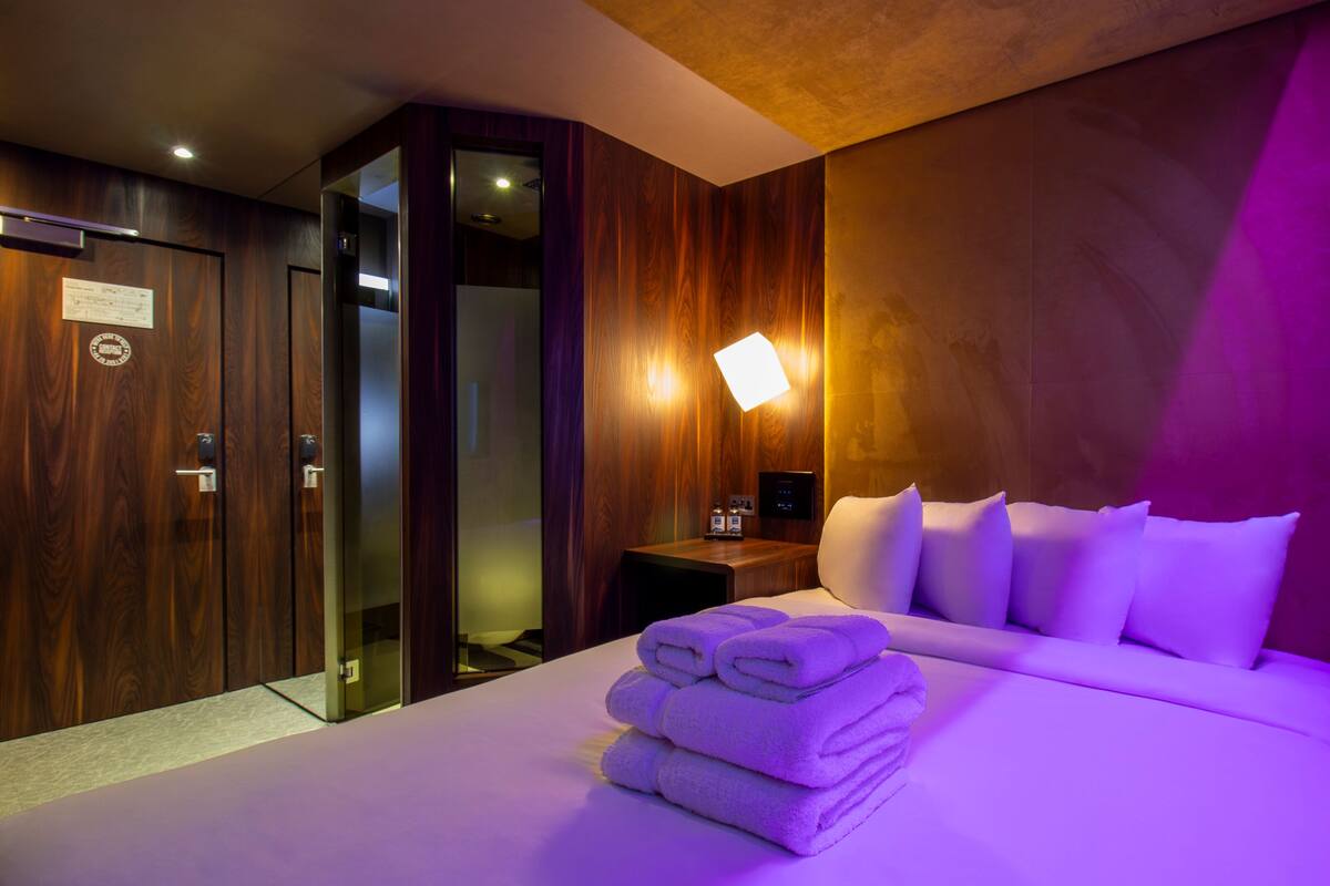 A hotel room with a neatly made bed, a wooden wardrobe, a nightstand with a lamp, and a bathroom visible through a glass door.