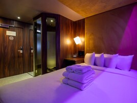 Habitación de hotel con una cama bien hecha, un armario de madera, una mesita de noche con lámpara y un baño visible a través de una puerta de cristal.