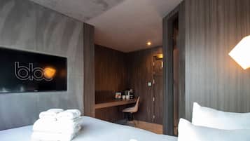 A modern hotel room with a bed, a desk, and a chair. There is a wall-mounted TV and a door leading to another room.