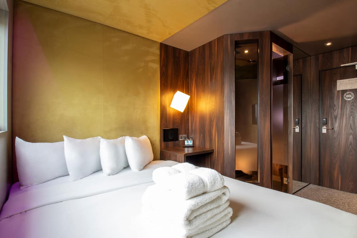 A hotel room with a large bed, a bedside table, a nightstand, a wall-mounted lamp, and a wooden wardrobe.