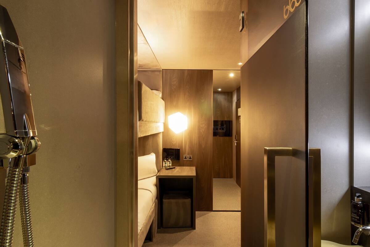 A hotel room with a wooden door, a bedside table with a lamp, a bed, and a small seating area.