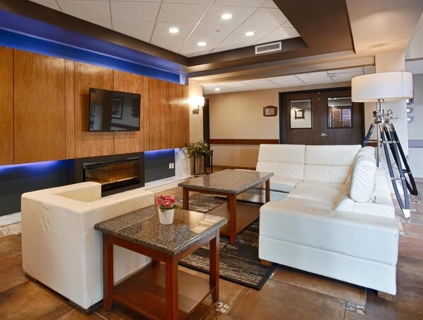 A modern hotel lobby with a white sofa, a small table with a vase of flowers, a flat-screen TV mounted on the wall, and a fireplace. - Days Inn by Wyndham Redwater (Redwater)