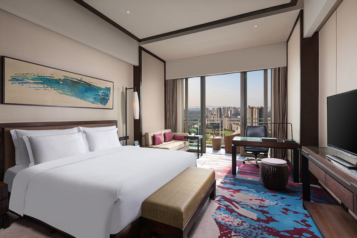A hotel room with a large bed, a desk with a chair, a television, and a view of the city through large windows.