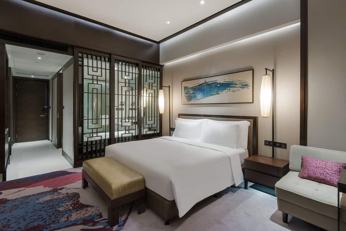 A modern hotel room with a large bed, a bench, a sofa, a decorative screen, and a painting on the wall.