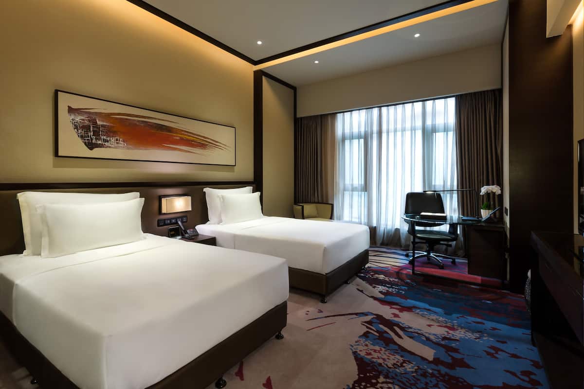 A hotel room with two beds, a desk, a chair, a large window with curtains, and a painting on the wall.