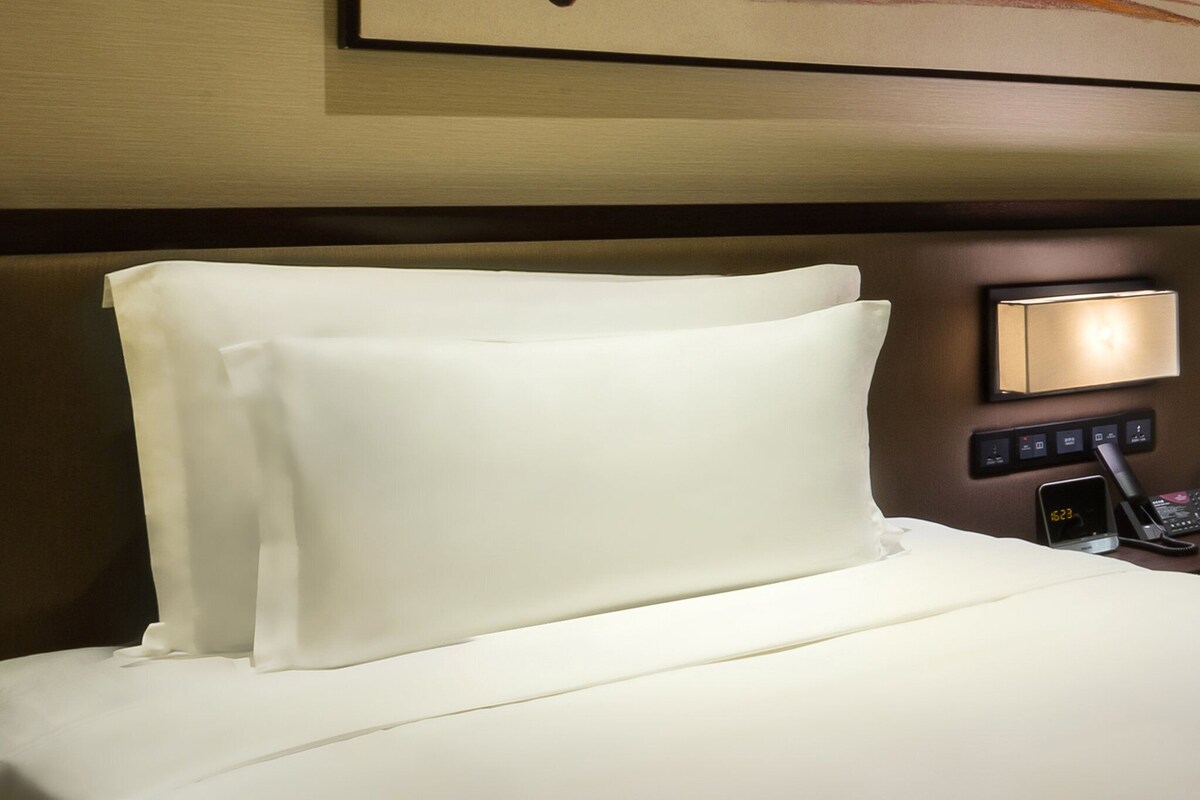 Una habitación de hotel con una cama bien tendida, una mesita de noche con una lámpara y un reloj digital que muestra la hora.